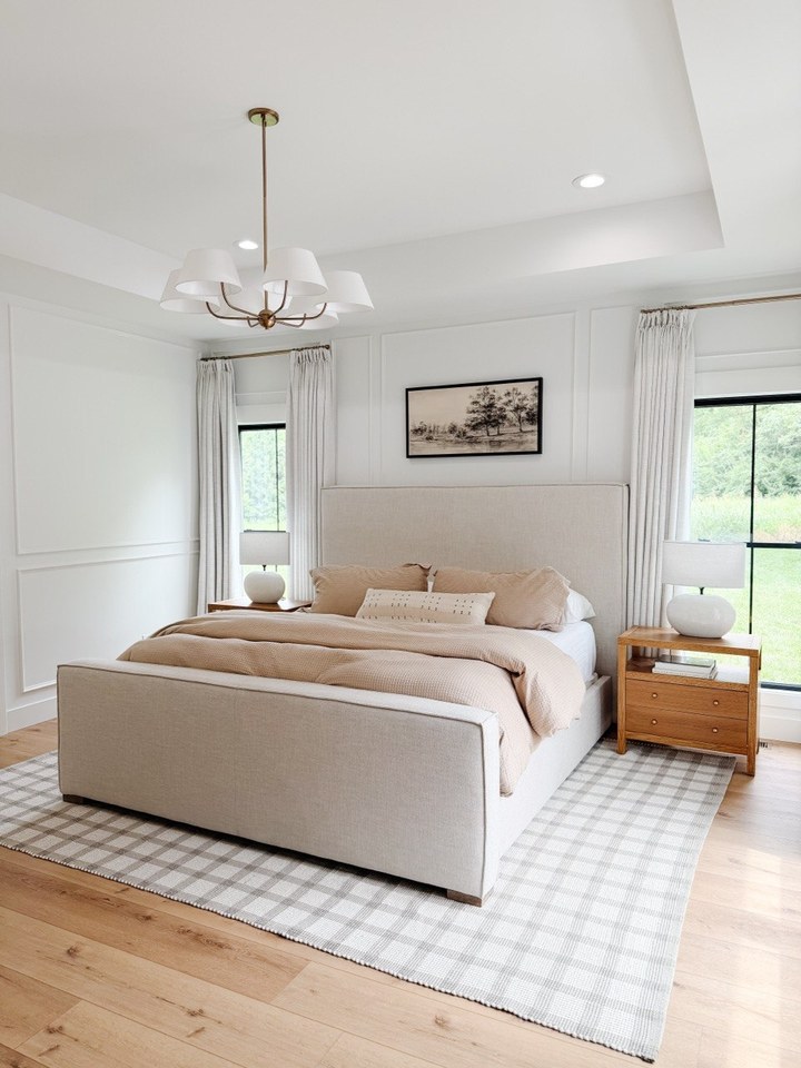 Neutral bedroom with an upholstered bed, wood nightstands, white lamps, plaid rug, paneled walls, and brass chandelier